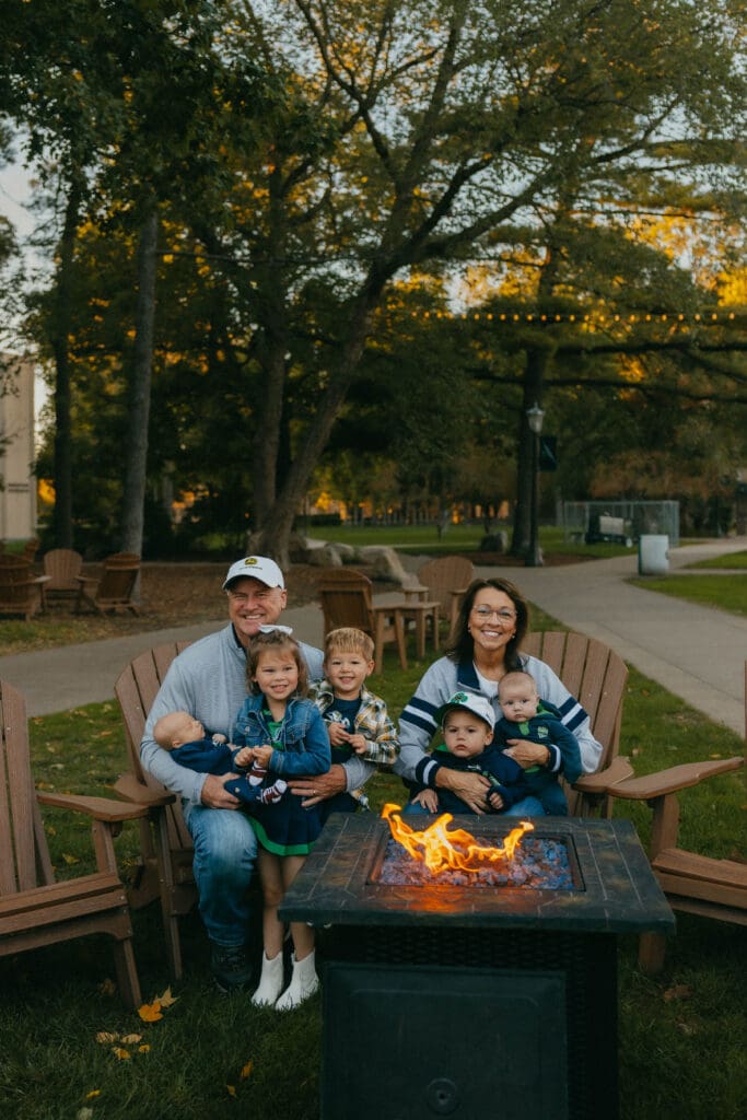 Grandparents cuddling their five grandchildren in outdoor chairs beside a fire pit.